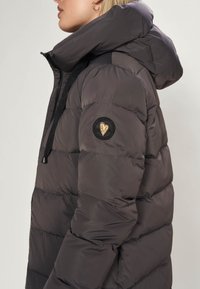 Donzen jas in donkergrijs met een hoge kraag en capuchon. Voorzien van doorgestikte panelen, elastische trekkoorden en een zwarte ronde patch met een gouden hart.