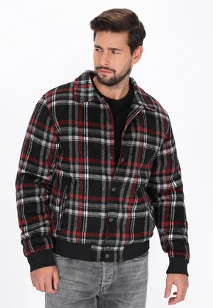 Bărbat cu păr închis la culoare și barbă, purtând o jachetă neagră, roșie și albă în carouri peste un tricou negru și blugi gri, uitându-se spre stânga.