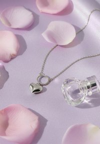 Sølvhalskæde med hjertemedaljon og snoet ringdetalje; lagt på blød lilla satin med pinke rosenblade og en glasparfume flaske.