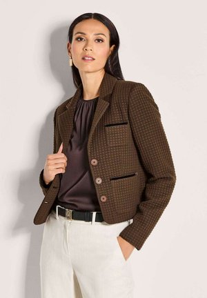 Vrouw in een bruine gestructureerde blazer, een donkerbruine satijnen blouse, witte broek en een zwarte riem met een "M" gesp, poserend tegen een lichte muur.