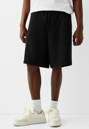 Schwarze Baumwollshorts mit elastischem Bund und Kordelzug, mit lockerem Schnitt und Saum, der bis zur Mitte des Oberschenkels reicht. Kombiniert mit weißen Turnschuhen.