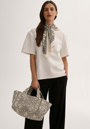 Kobieta ubrana w biały top z krótkim rękawem i czarne spodnie, trzyma dużą torebkę z wzorem, z dopasowaną chustą z tym samym wzorem związaną wokół szyi.