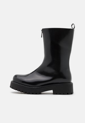 Botte en cuir noire brillante mi-mollet avec semelle épaisse rainurée, fermeture éclair latérale et languette sur fond blanc.