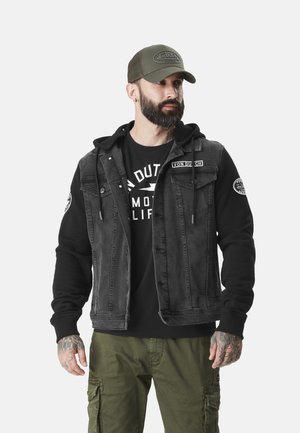Skægget mand iført grøn Von Dutch-kasket, sort hoodie med denimvest, sort grafik t-shirt og olivengrønne cargobukser, står og kigger til højre.