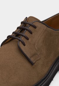 Καφέ σουέτ παπούτσι με κορδόνια και σκούρες καφέ λεπτομέρειες, με ραμμένη διακόσμηση και μαύρη καουτσούκ σόλα.