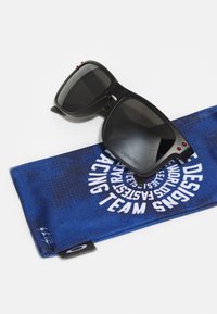 Sorte solbriller med mørke linser, røde detaljer og mat finish, vist på en blå rengjøringsklut med hvit tekst og mønstre.