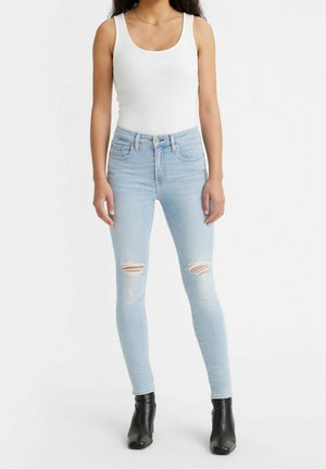Frau trägt hellblaue zerrissene Skinny-Jeans, weißes ärmelloses Oberteil, schwarze Knöchelstiefel, steht vor einem schlichten weißen Hintergrund.