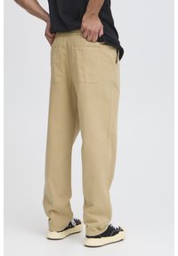 Pantaloni beige, ampi, con vita elasticizzata, due tasche posteriori e una texture liscia. Indossati con sneakers nere e bianche.