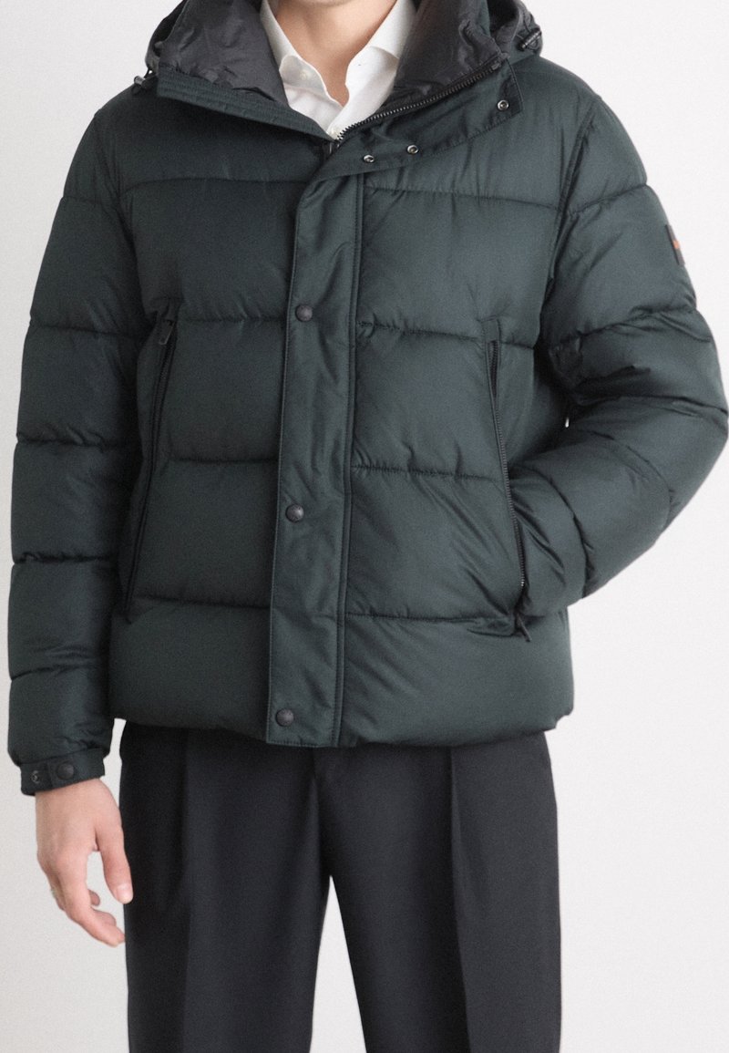 Uomo che indossa un giubbotto imbottito verde scuro sopra una camicia bianca e pantaloni neri, con una mano in tasca, su uno sfondo neutro.