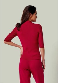 Rotes geripptes Strickoberteil mit drei Viertelärmeln, das einen hohen Kragen und eine figurbetonte Silhouette aufweist, kombiniert mit passenden roten Hosen.