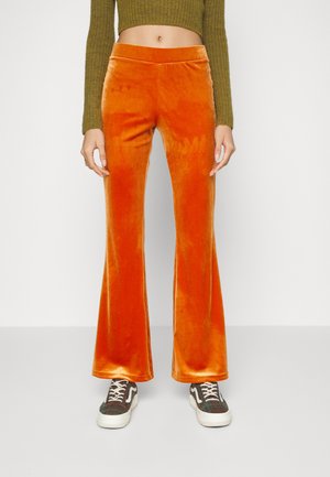 Oranssi samettihousut, joissa on leveät lahkeet ja sileä pinta, korkea vyötärö ja pehmeä kiilto, yhdistettyinä mustavalkoisiin kuviollisiin lenkkareihin.