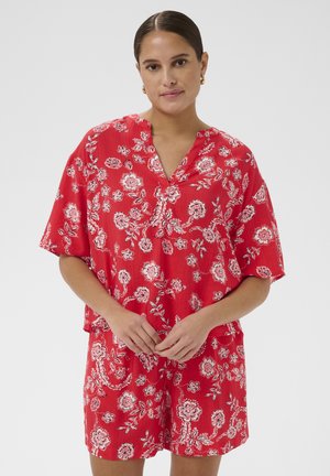 Femme aux cheveux lissés portait un chemisier rouge à manches courtes à motifs floraux et un short assorti, debout devant un fond clair uni.