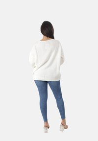 Witte gebreide oversized trui met een geribde onderrand, gecombineerd met blauwe skinny jeans. De achterkant toont een ontspannen pasvorm en een zachte textuur.