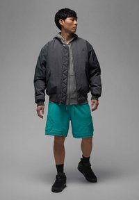 Grå bomberjakke over en grå hættetrøje, parret med teal-shorts. Modellen har sorte løbesko og sorte ankelstrømper på.