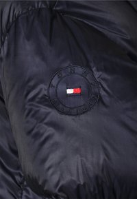 Marineblå puffjakke med et rundt broderet logo i røde, hvide og blå nuancer. Glat, skinnende stof med kviltet tekstur.