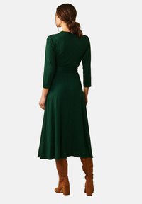 En kvinna står med ryggen mot betraktaren, klädd i en lång, grön klänning och bruna högklackade stövlar, mot en enkel vit bakgrund.