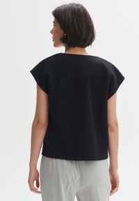 Zwarte, gestructureerde top met een losse, vierkante pasvorm en korte mouwen, gedragen over lichtgrijze broeken. Eenvoudig en minimalistisch ontwerp.