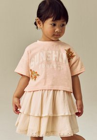 Rosa Kurzarm-T-Shirt mit dem Aufdruck "SUNSHINE FOREVER" und floraler Stickerei, kombiniert mit einem geschichteten cremfarbenen Rock mit Spitzendetails.