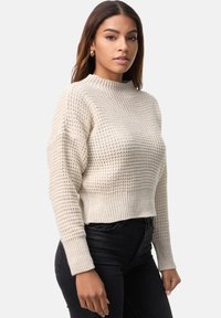 Sweter wykonany z dzianiny w jasnym beżu, z teksturowanym wzorem, wysokim kołnierzem i skróconym krojem, zestawiony z ciemnymi dżinsami. Ma ściągacze na rękawach i u dołu.