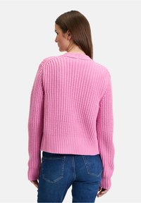 Helles rosa gestricktes Sweatshirt mit geripptem Saum und Bündchen, das ein strukturiertes Muster aufweist. Über dunkelb blauen Jeans getragen, was einen lässigen Look zeigt.