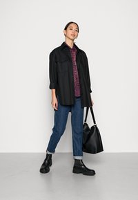 Camisa preta de botões sobre uma blusa floral roxa, jeans azuis com as bainhas dobradas e botins pretos chunky. Bolsa preta pendurada no braço.