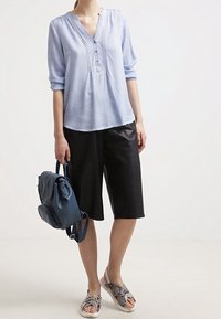 Kobieta nosząca jasnoniebieską bluzkę z podwiniętymi rękawami, czarne skórzane szorty do kolan, sandały w wężowy wzór, trzymająca granatowy plecak.