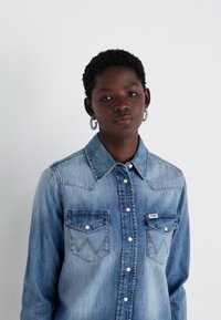 Camisa de denim azul com botões de pressão, dois bolsos no peito com costura em zigzag e colarinho pontudo. O tecido parece ligeiramente desbotado.