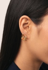 Boucles d'oreilles en or en forme de crabe avec une pierre précieuse orange, présentées sur des cheveux foncés et un fond neutre.