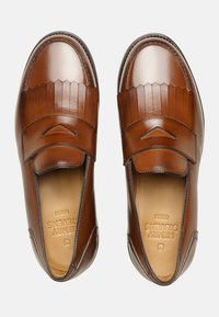 Scarpe in pelle marrone con punta rotonda, dettagli frangiati alzati e design slip-on con hardware minimo. Texture liscia su tutta la superficie.