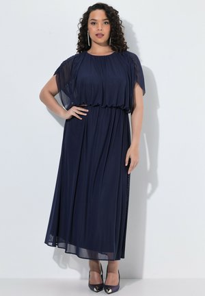 Femme aux cheveux bouclés portant une robe longue bleu marine avec des manches transparentes et des talons noirs pointus, debout contre un mur blanc.