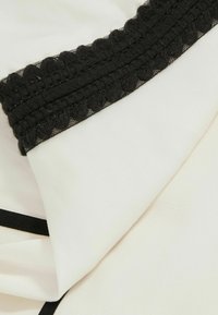Weißer Stoff mit einem strukturierten Bereich, der mit einer schwarzen Spitze und einem geflochtenen Design entlang des Randes kontrastiert, im Gegensatz zu einer glatten Oberfläche.