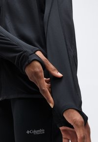 Κοντινό πλάνο ατόμου που ρυθμίζει την τρύπα για τον αντίχειρα σε μαύρο, υφασμάτινο μακρυμάνικο κορμάκι με ορατό λογότυπο Columbia Titanium σε μαύρο παντελόνι.