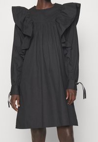 Sort knælængde kjole med lange pufærmer og store flæsedetaljer på skuldrene, båret af en stående person med mørk hudtone.