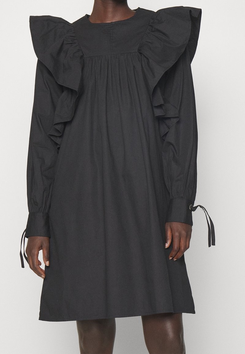 Sort knælængde kjole med lange pufærmer og store flæsedetaljer på skuldrene, båret af en stående person med mørk hudtone.