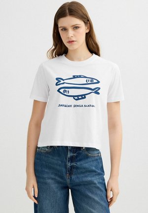 Joven mujer que lleva una camiseta blanca corta con ilustraciones azules de sardinas y texto en italiano, combinada con vaqueros azules de talle alto.