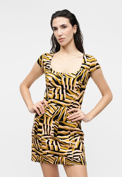 Γυναίκα με σκούρα μαλλιά, που φοράει κοντομάνικο φόρεμα με αφηρημένο κίτρινο και μαύρο σχέδιο, στέκεται με τα χέρια στη μέση μπροστά σε ένα απλό φόντο.