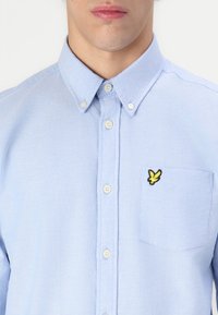 Chemise boutonnée en coton bleu clair avec une poche poitrine gauche et un logo brodé jaune. Les boutons sont de couleur crème, avec un col classique.