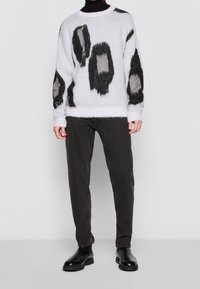 Weißer, flauschiger Pullover mit abstrakten Mustern in Schwarz und Grau, mit gerippten Bündchen und Saum, kombiniert mit schwarzen, schmal geschnittenen Hosen und schwarzen Stiefeln.