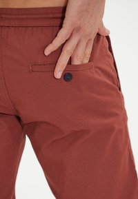Rostfärgade shorts i lätt tyg, med en bakficka som har en svart knapputstyrsel och ett elastiskt midjeband.
