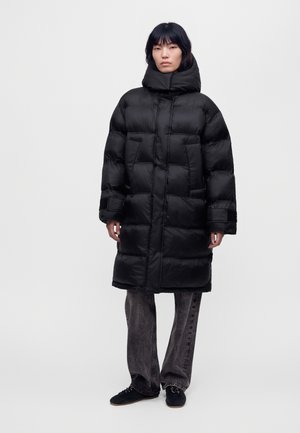 Une personne porte un long manteau matelassé noir à col montant, un jean gris ample et des chaussures noires sur un fond clair uni.