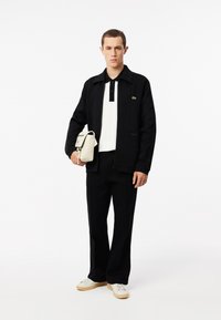 Chaqueta negra con cremallera sobre una camiseta polo blanca, combinada con pantalones negros y zapatillas blancas. El modelo sostiene un pequeño bolso color crema.