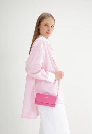 Rosa crossbody taske med guldkæde-rem; glat tekstur, rektangulær form og logo-detalje. Modellen bærer en pink stribet skjorte.