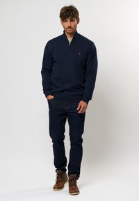 Marinefarbener Reißverschluss-Pullover mit gerippten Bündchen, kombiniert mit dunklen Jeans und braunen Schnürstiefeln. Mit kleinem rotem Logo auf der Brust.
