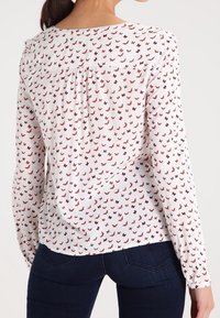 Vit blus med ett fjärilsprint i röd och mörkrosa, långa ärmar, en ihopdragen bakrygg och en rundad nederkant. Bärs med mörkblå jeans.