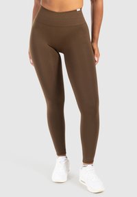 Donna che indossa collant marroni a vita alta e sneakers bianche, in piedi contro uno sfondo grigio chiaro uniforme.