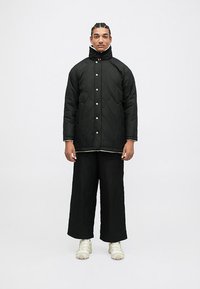 Giacca nera trapuntata con colletto alto, bottoni a pressione e pantaloni ampi abbinati. Indossata con scarpe sportive di colore chiaro.