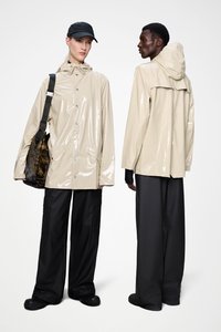 Μπεζ γυαλιστερό αδιάβροχο με κουκούλα, διαθέτει κουμπώματα με κουμπιά, πλαϊνές τσέπες και άνετη γραμμή, συνδυασμένο με μαύρο παντελόνι φαρδιάς γραμμής και μαύρα παπούτσια.