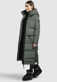 Cappotto lungo imbottito verde con colletto alto, chiusura con zip e tasche laterali, abbinato a stivali neri con finitura testurizzata.
