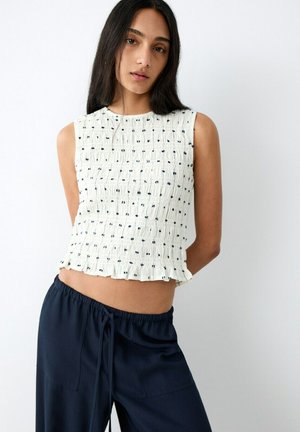 Femme aux longs cheveux noirs portant un crop top blanc texturé sans manches avec de petits points noirs et un pantalon à cordon bleu marine.