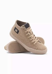 Zapatillas altas de color beige hechas de tela con un acabado texturizado, que presentan un acento de cuero oscuro, puntera redonda y una suela de goma con patrones de agarre.